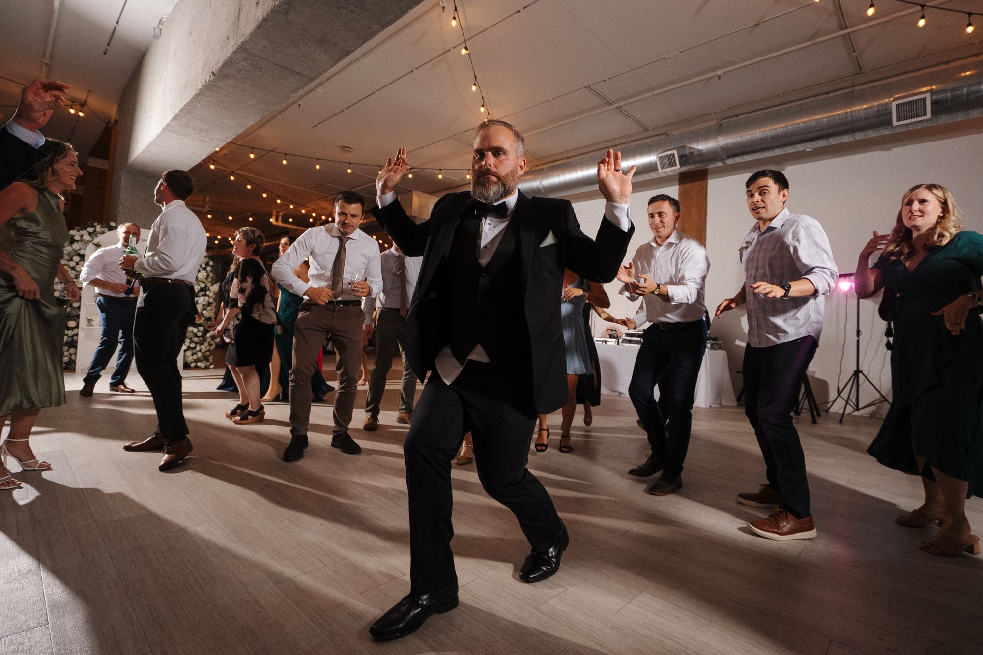 Dancers twirl and move across a wooden floor during a ballroom dance event with string lights overhead.