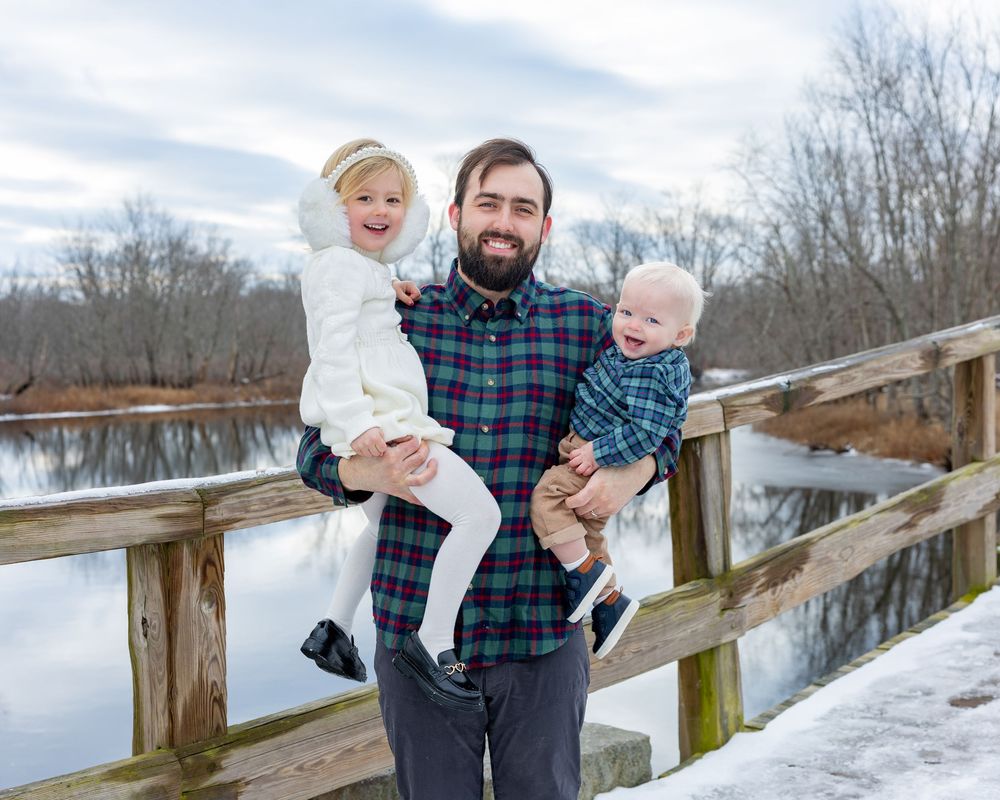 Father and children pose on Old North Bridge during a bright afternoon during winter family portrait session in Concord, MA.