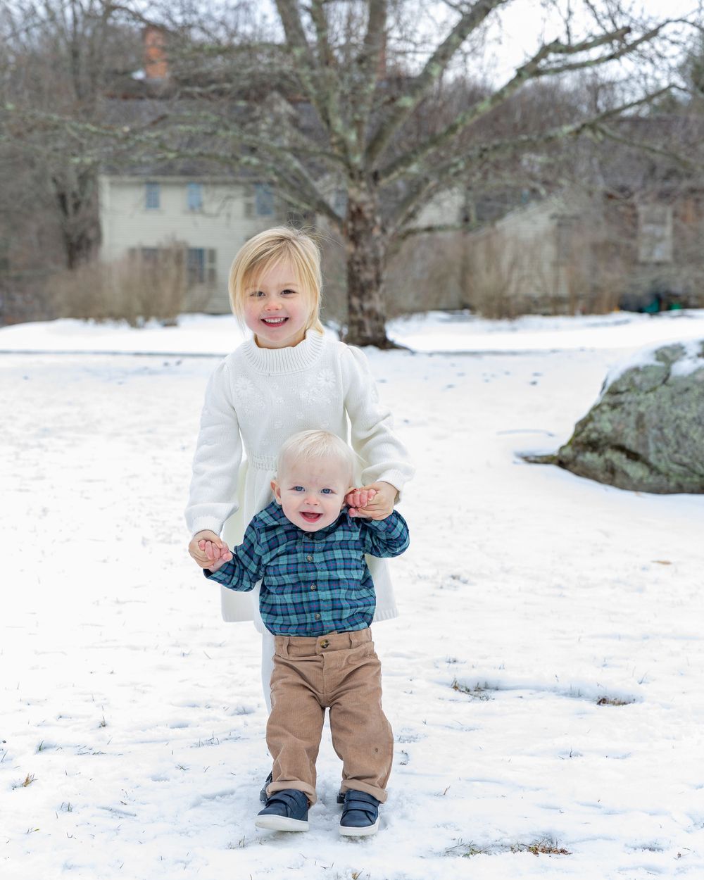 Two children playing together in a snowy winter landscape with a historic house in Concord, MA during professional family photo session.