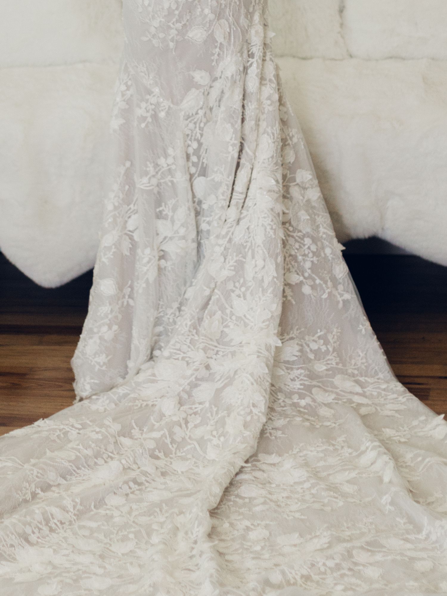 Close-up view of a white lace wedding dress train with intricate floral embroidery details spread across wooden flooring.