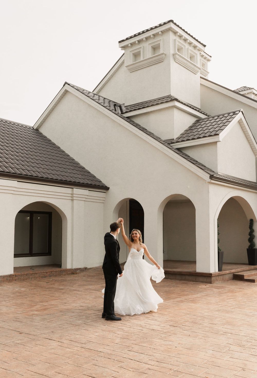 A couple dancing on a terrace in front of a white church-style building with arched walkways and terra cotta tile flooring.