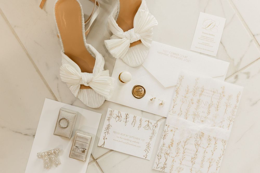 Elegant wedding accessories including white bow shoes, rings, and handwritten invitation cards arranged on white surface.