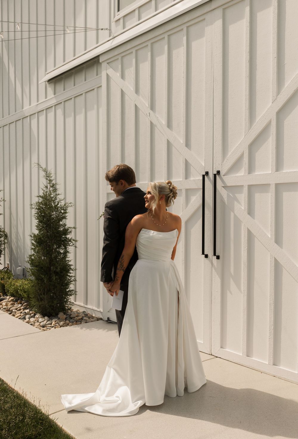 Couple in wedding attire standing outside white barn doors with evergreen tree against rustic white paneled wall.