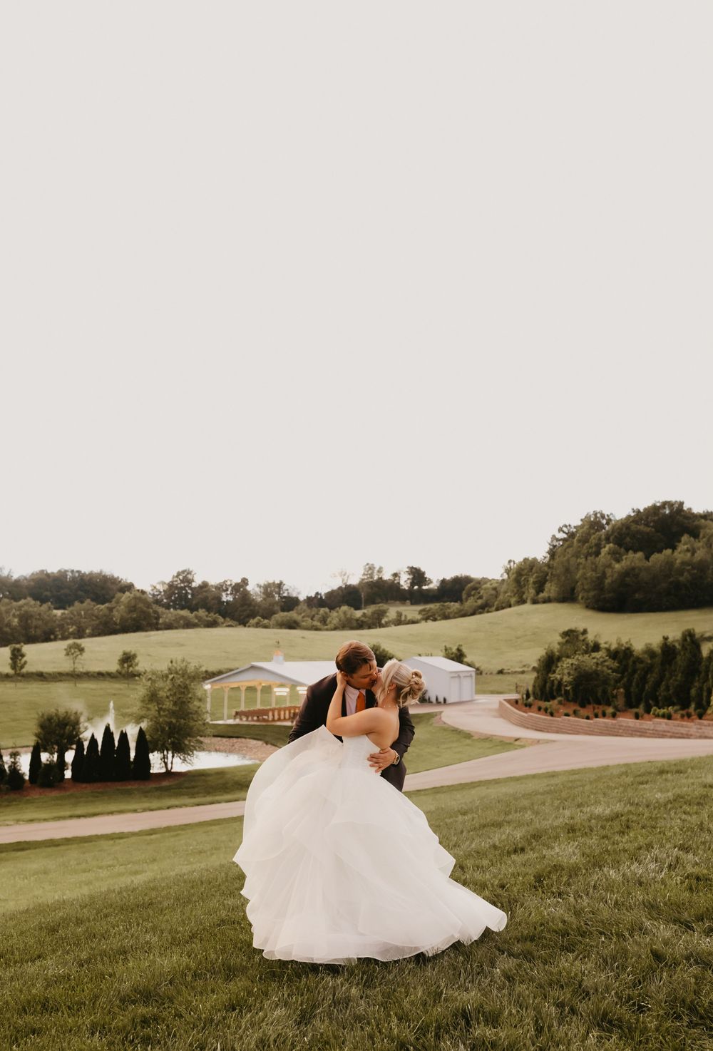 A couple sharing a romantic moment on a grassy hillside overlooking a rustic venue with green rolling hills in the distance.