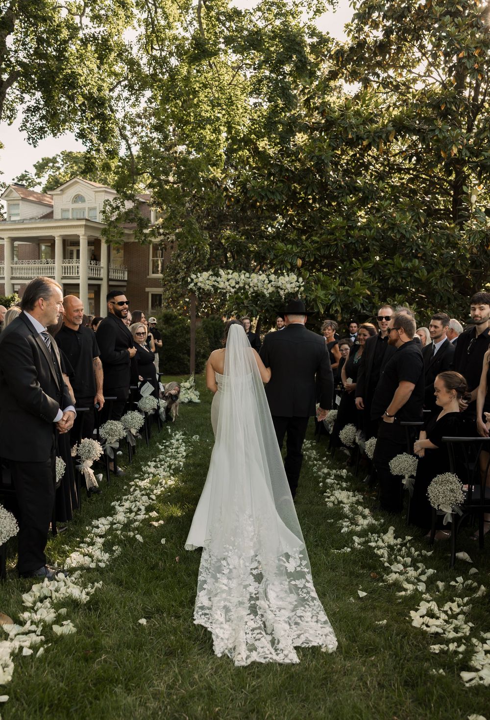 An outdoor wedding ceremony setup features a scenic aisle lined with delicate white floral arrangements and green foliage.
