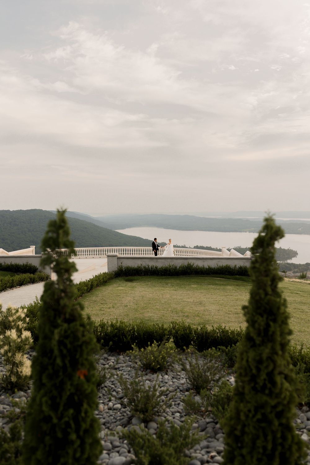 A scenic overlook with cypress trees framing a view of mountains and lake through a white railing on a misty day.
