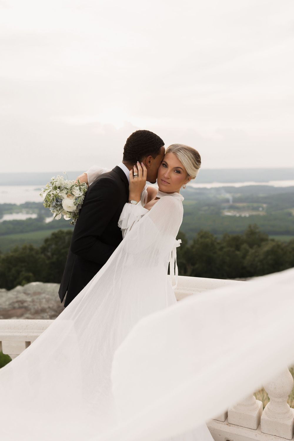 Romantic wedding couple embraces on balcony overlooking scenic landscape as bride's white dress flows in breeze.