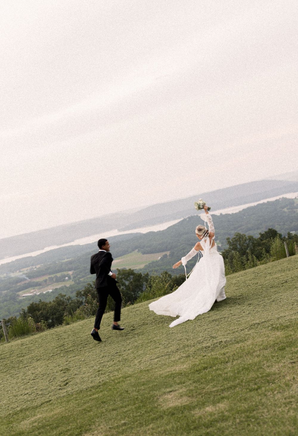 A couple in wedding attire playfully run across a grassy hilltop with mountains and valleys in the scenic background.