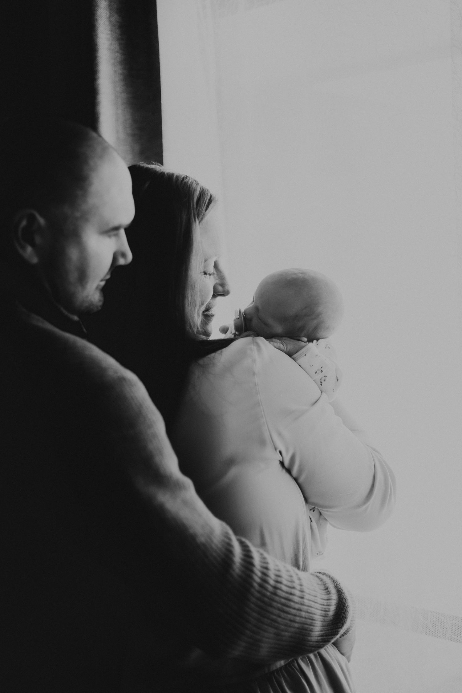 A couple stands by a window in silhouette holding their newborn baby in a tender black and white moment.