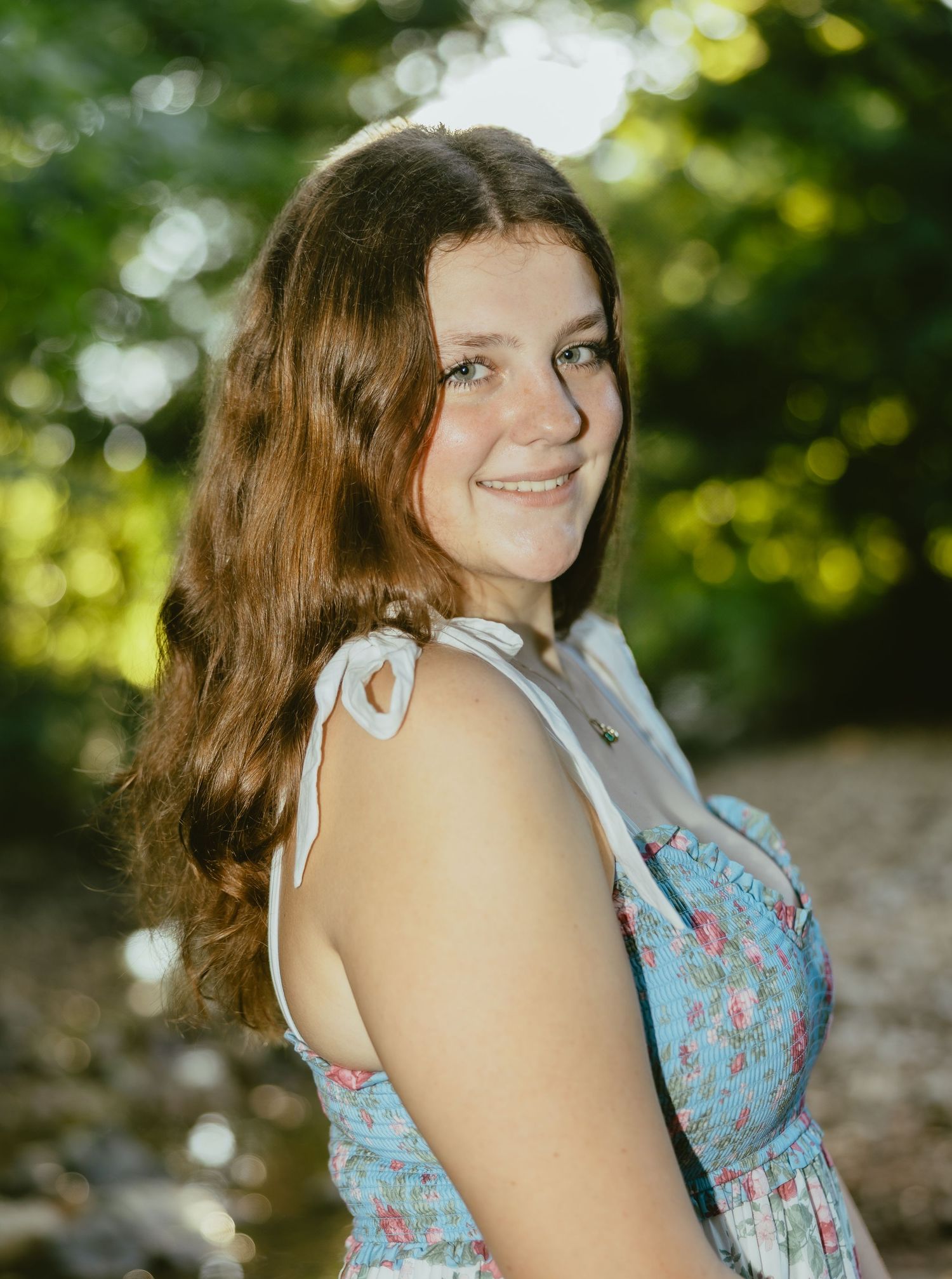 Woman in blue floral summer dress poses outdoors in dappled sunlight with natural bokeh effect in background.