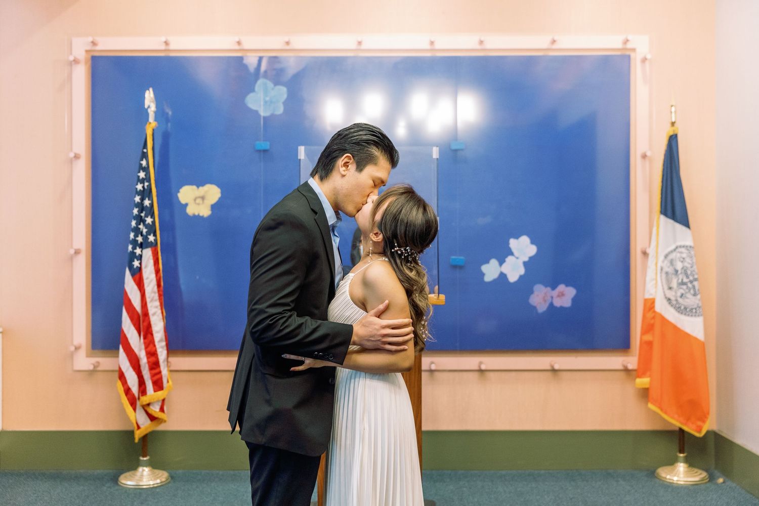 A couple shares a romantic kiss in front of American and NYC flags in a city hall ceremony room.