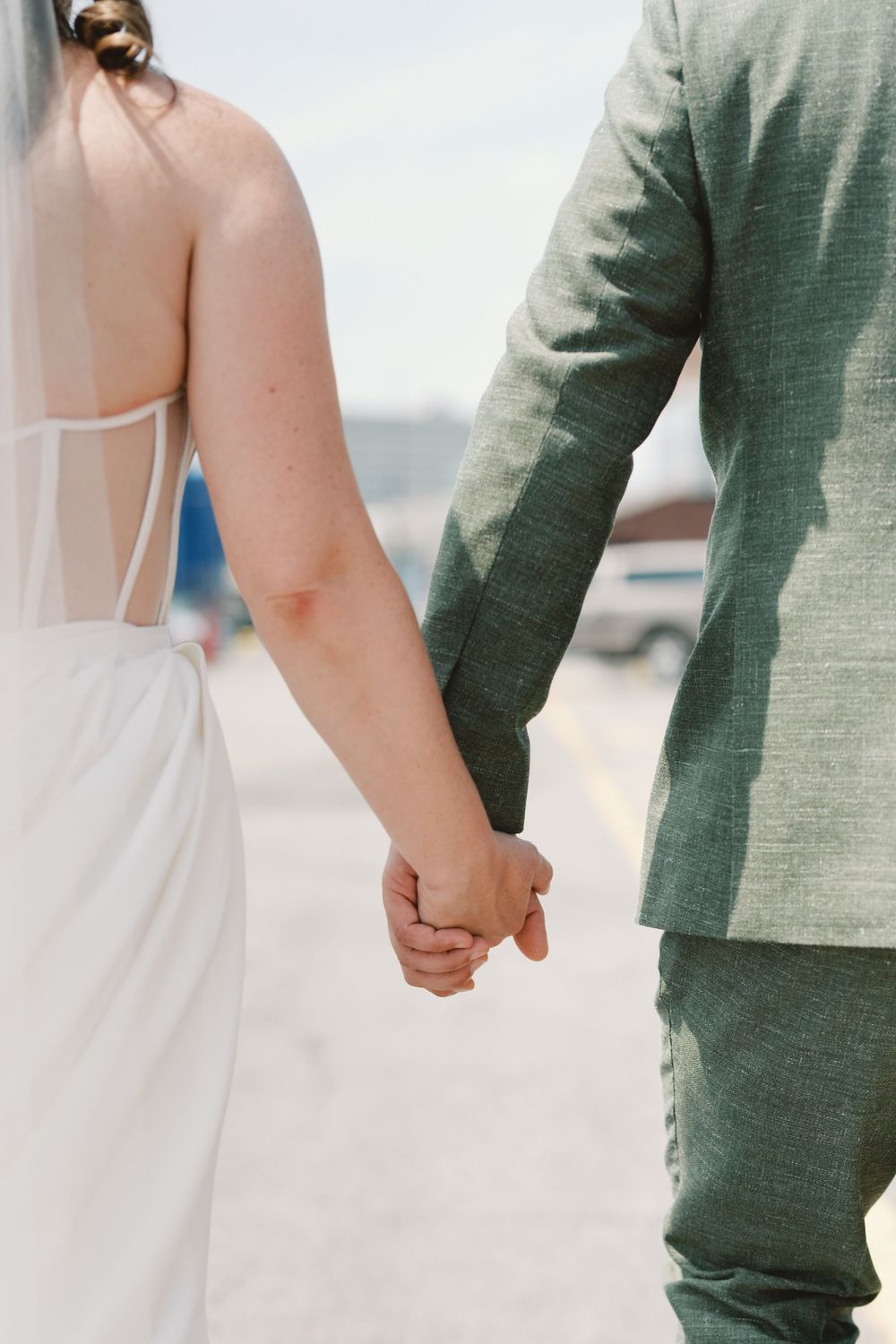 Close-up view of a couple holding hands, with bride in white dress and groom in sage green suit on their wedding day.