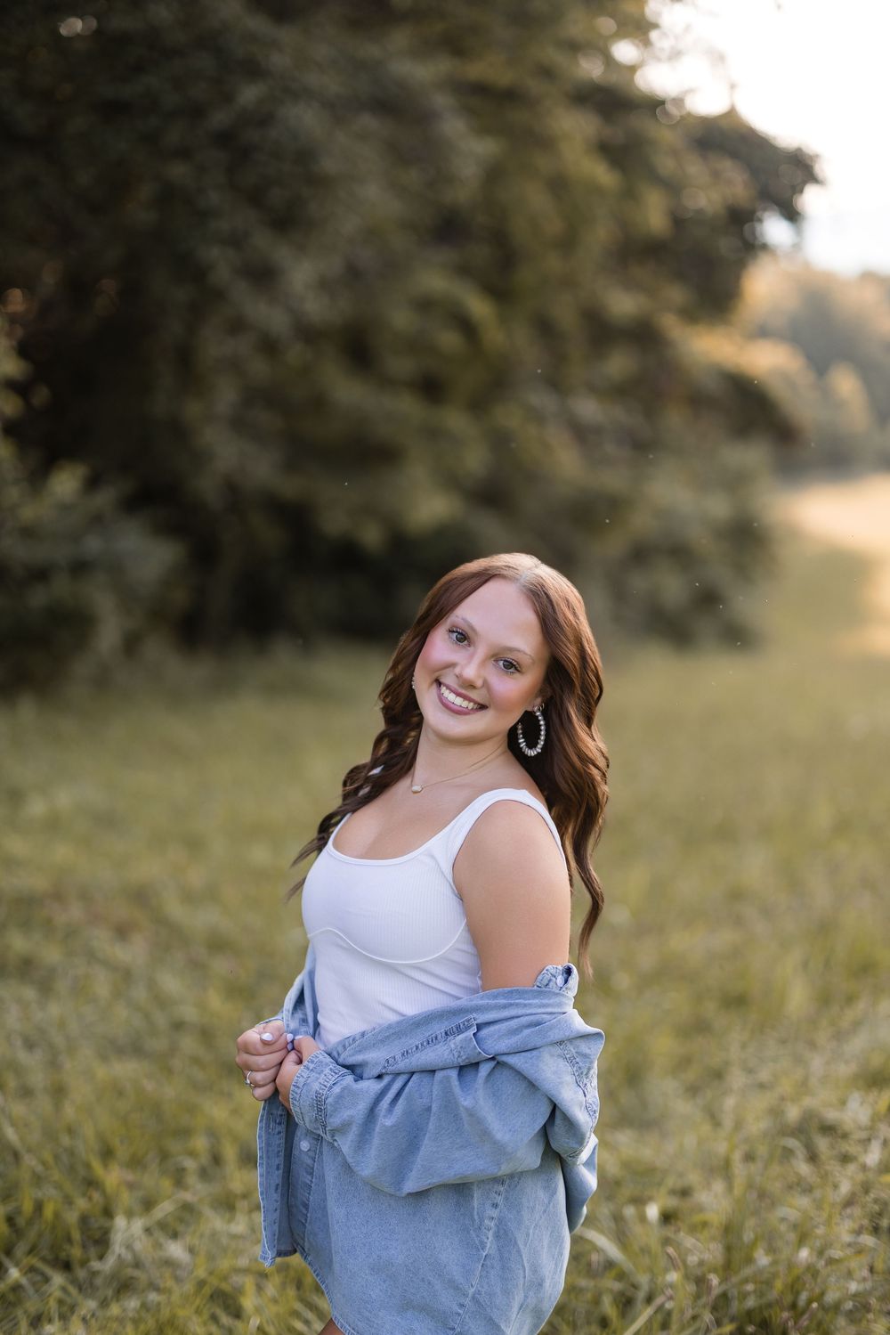 Casual outdoor portrait of a smiling young woman in a white tank top and denim jacket in a grassy field at sunset.
