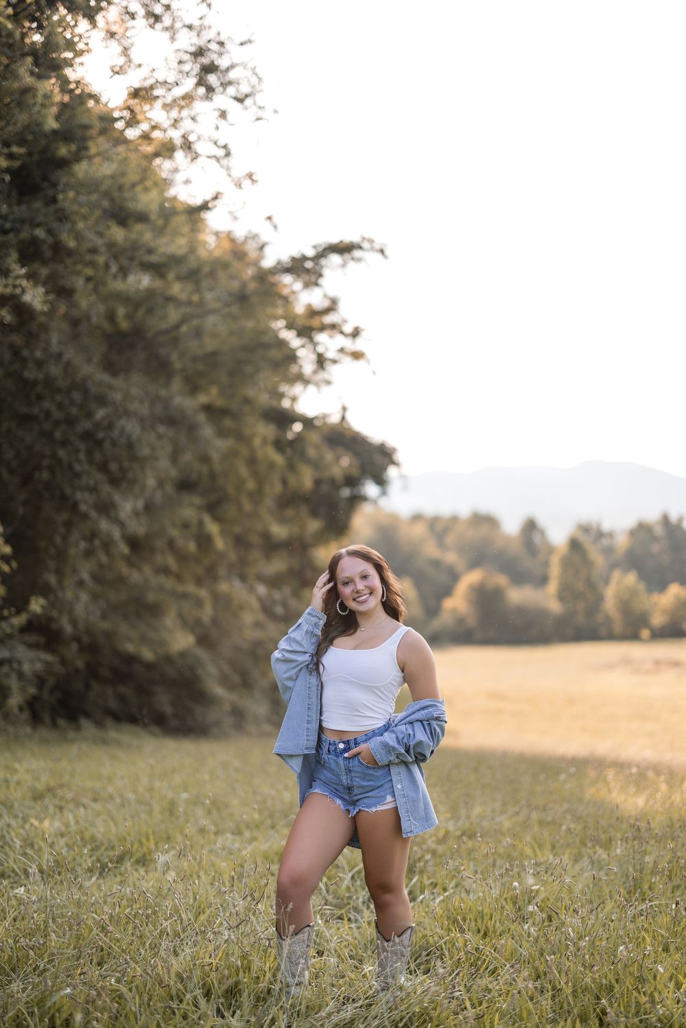 Person in denim shorts and white top poses outdoors in a sunlit meadow with mountains and autumn trees in background.