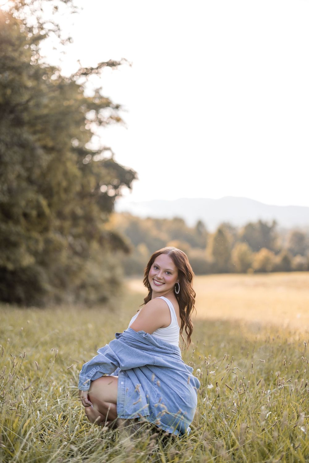 A figure in a denim dress sits gracefully in a golden meadow during sunset with trees in the background.