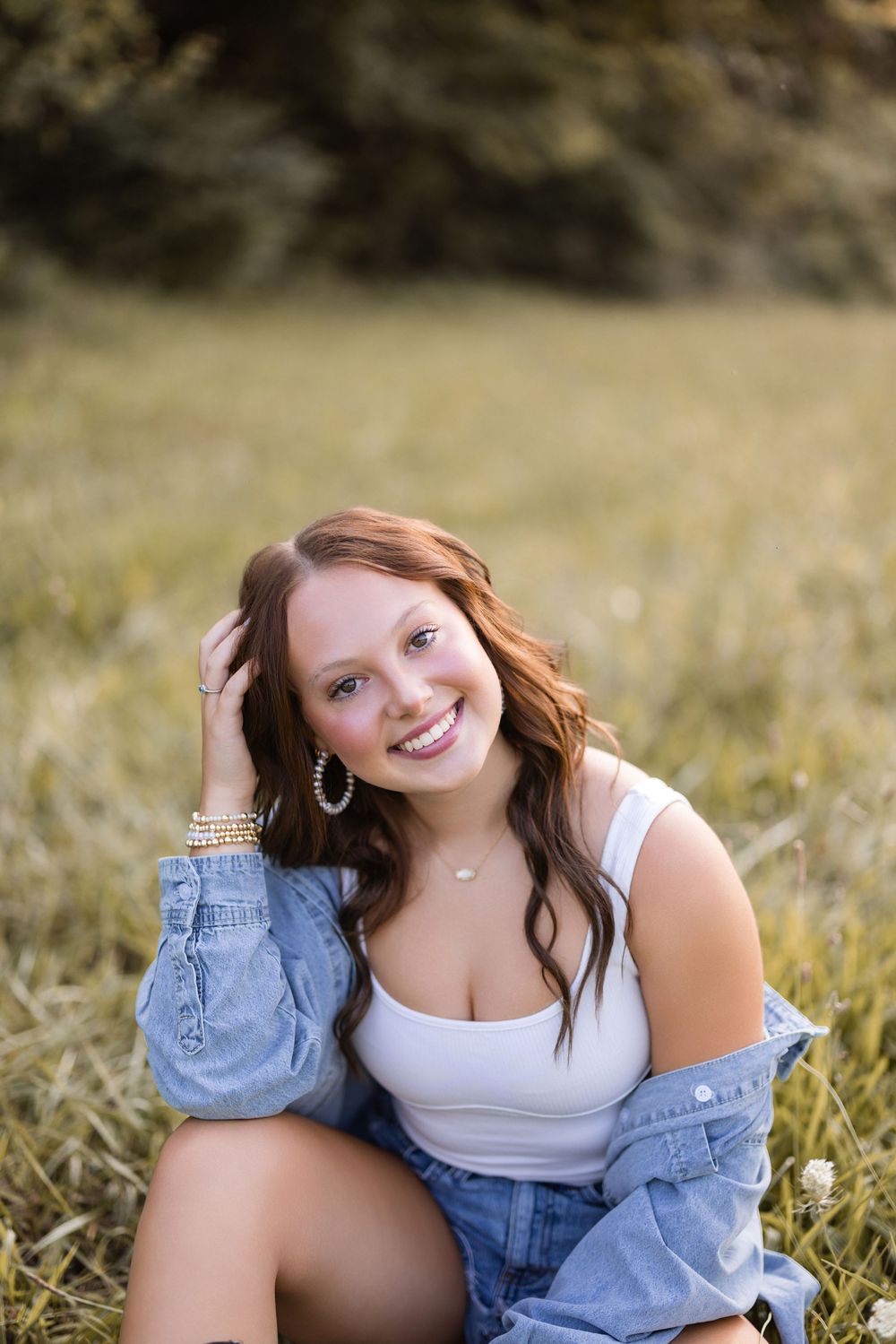 Person wearing a white tank top and denim jacket sits in golden grass during an outdoor portrait session.