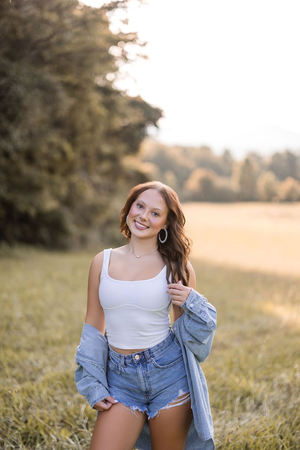Casual summer fashion portrait in a meadow with denim jacket and white tank top at sunset.