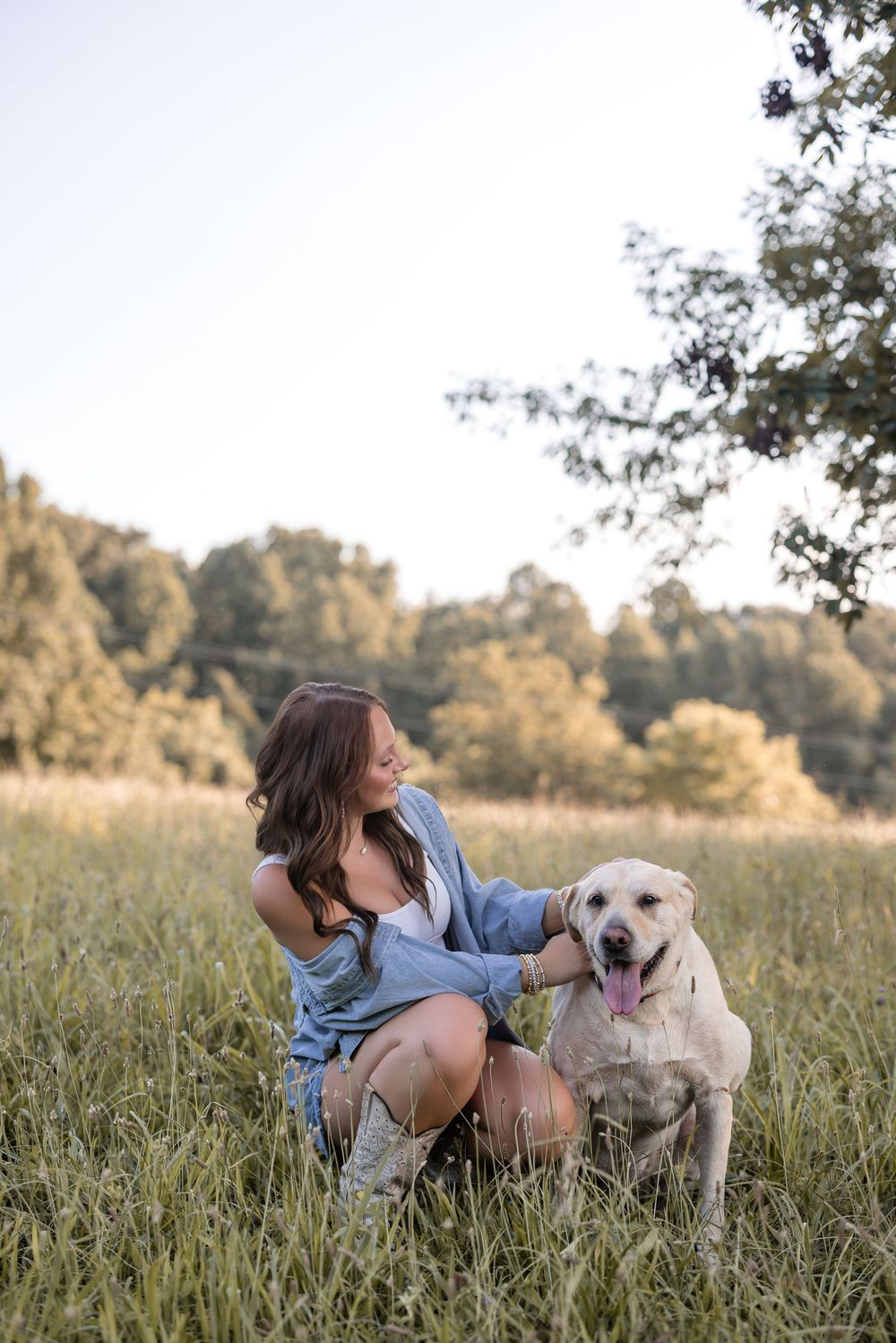 A woman in denim sits with her happy Labrador retriever dog in a golden summer meadow at sunset.