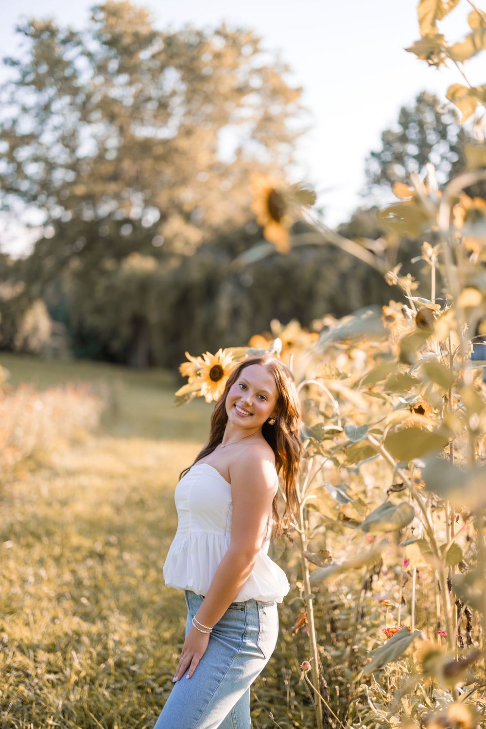 A joyful summer portrait among tall sunflowers in warm evening light wearing a white top and jeans.