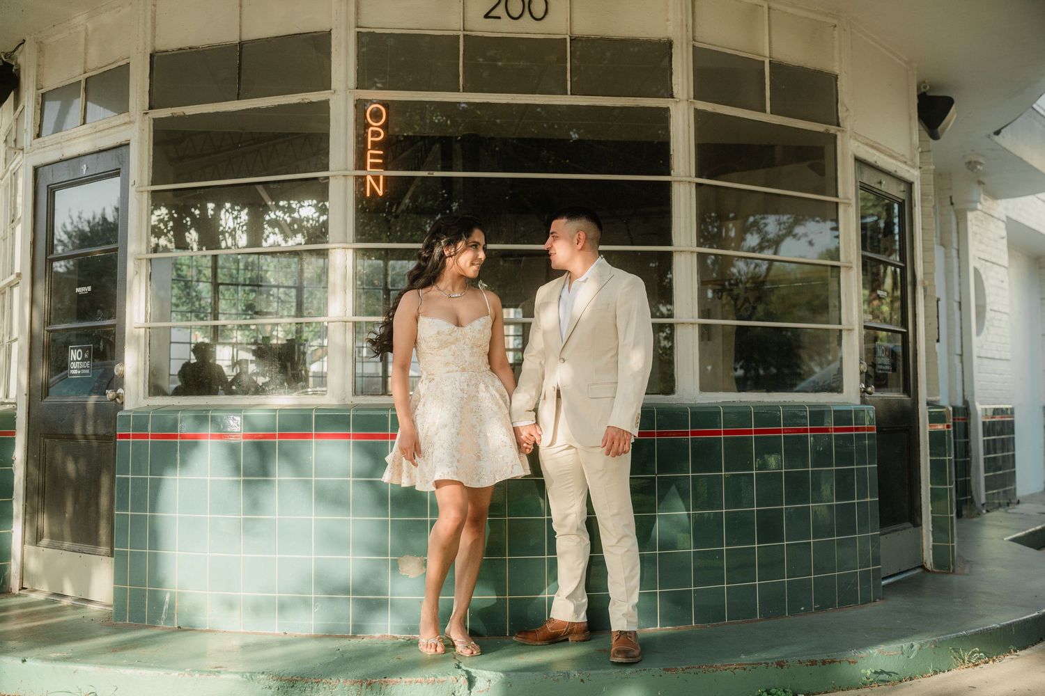 A couple walks together along a sidewalk with palm trees and Spanish-style architecture in the background.