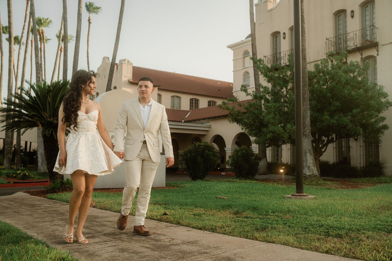 A romantic couple walking on a path lined with palm trees near a Mediterranean-style building.