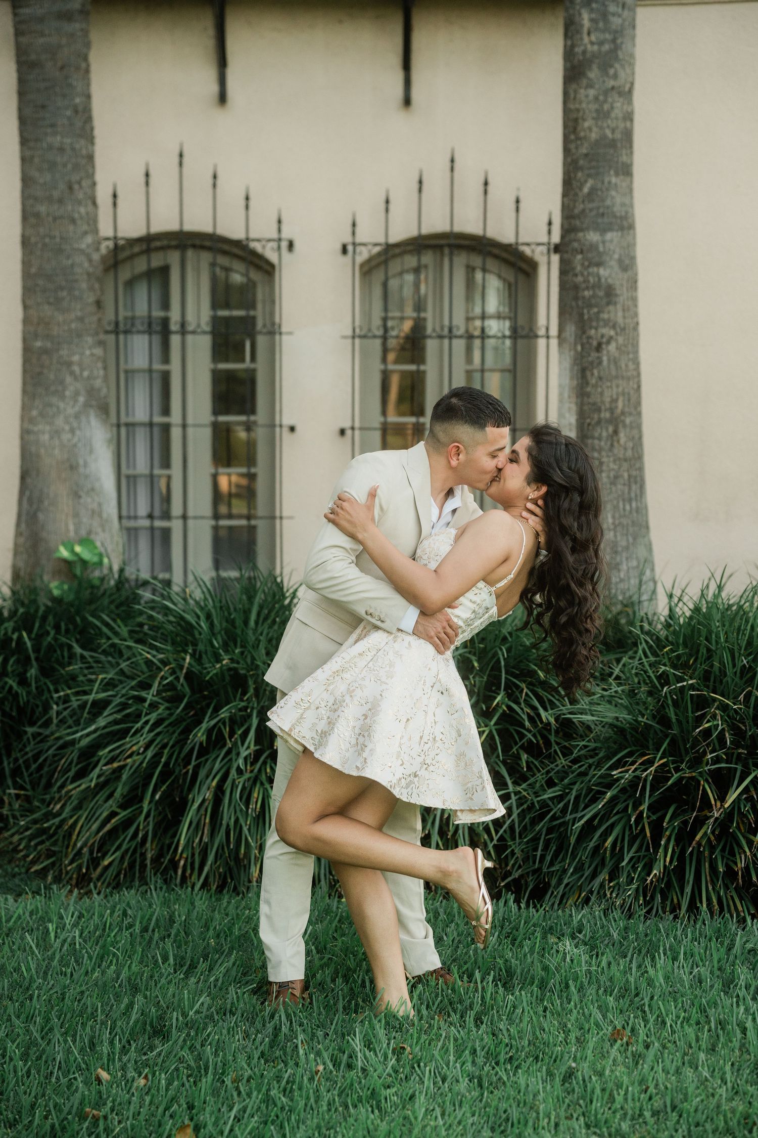A romantic moment shared on a grassy lawn in front of a Spanish colonial building with palm trees.