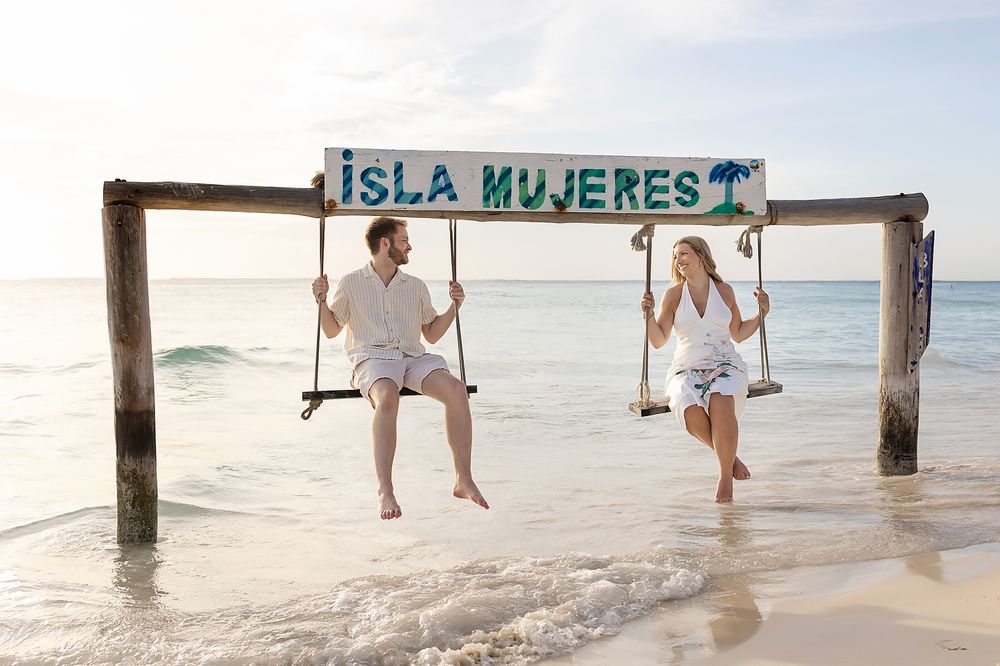 Two people on swings under Isla Mujeres sign at a tropical beach with white sand and turquoise water.