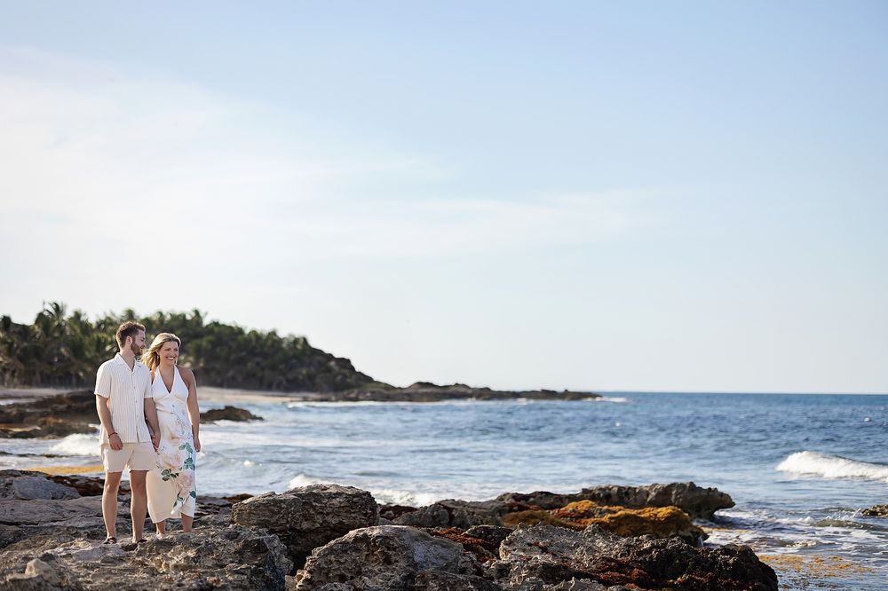 Two people in white outfits walk along a rocky beach coastline with waves crashing against the shore on a sunny day.