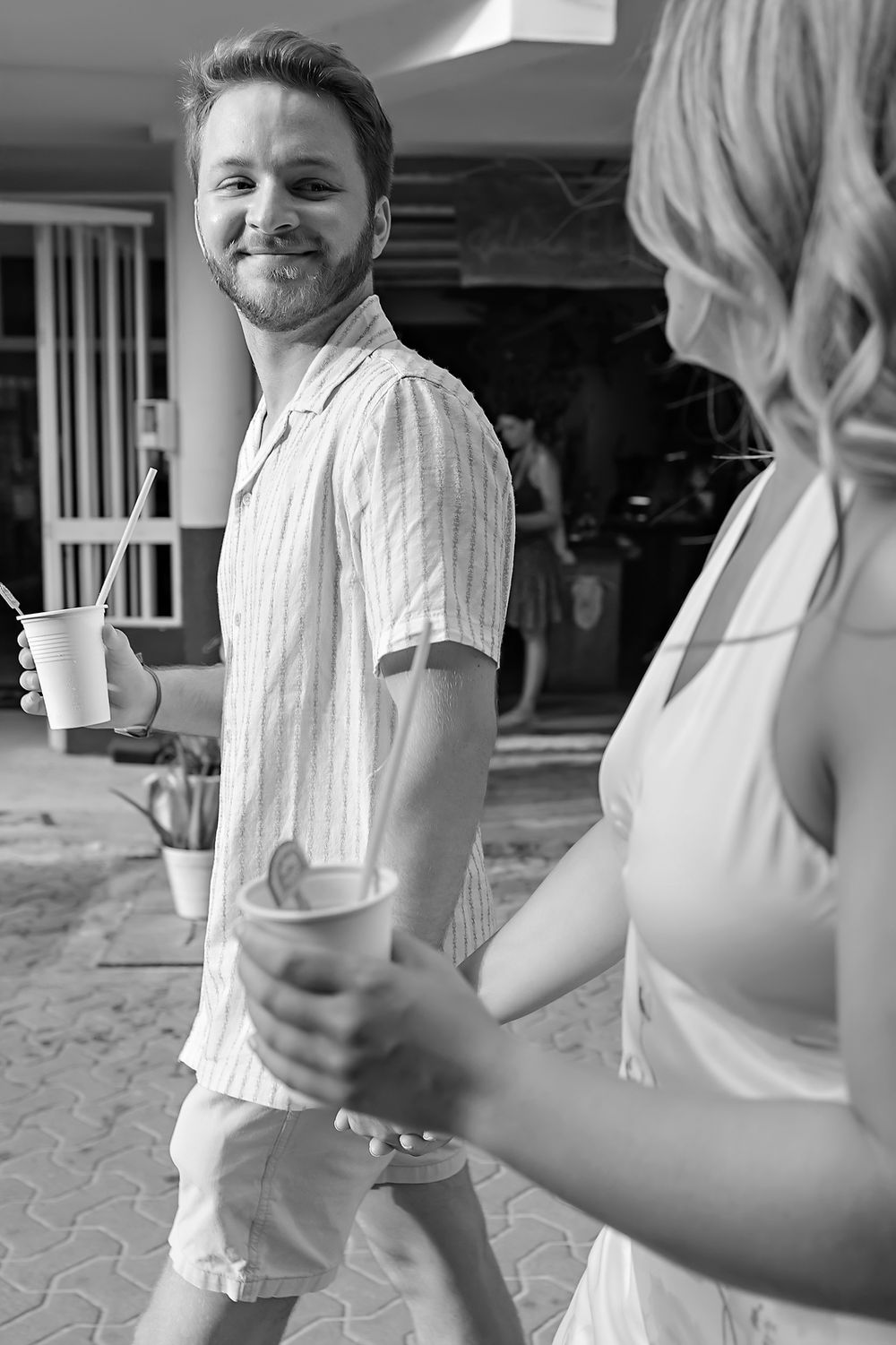People handing coffee cups back and forth in a casual outdoor setting while wearing light-colored clothing.