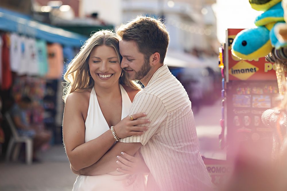A couple shares a romantic moment laughing together at a carnival with colorful blurred attractions in the background.