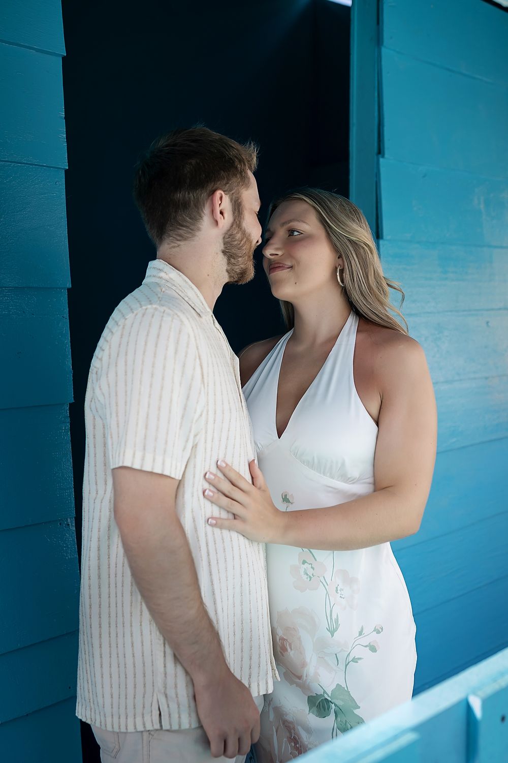 A romantic couple shares an intimate moment against a vibrant blue wall, both wearing white outfits.