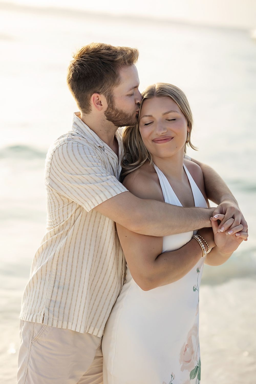 Romantic couple embraces on beach at sunset in light summer outfits sharing tender moment together.
