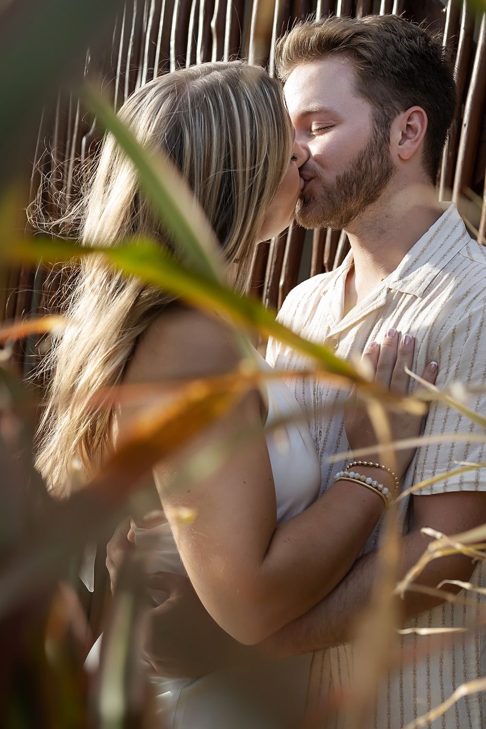 Romantic couple embracing and kissing in soft natural light with blurred foliage in foreground.