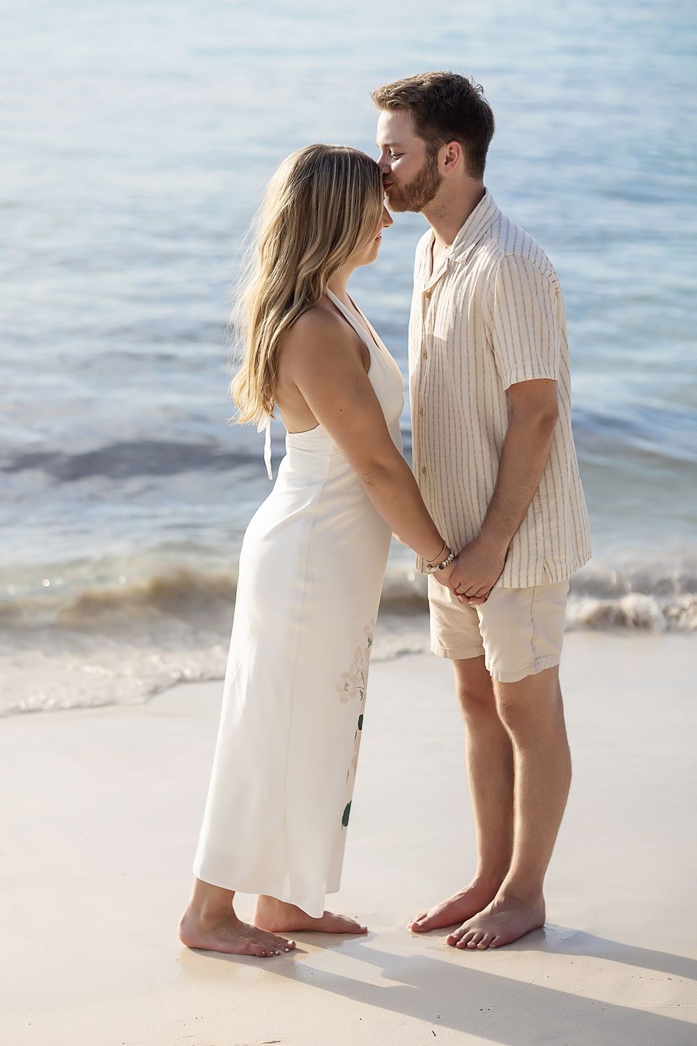 A romantic couple in light summer attire sharing an intimate moment while standing barefoot on a beach at sunset.
