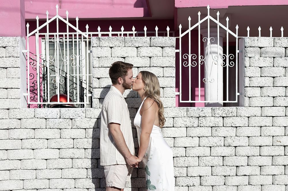 A couple in white outfits share an intimate moment against a white brick wall with decorative iron gates.