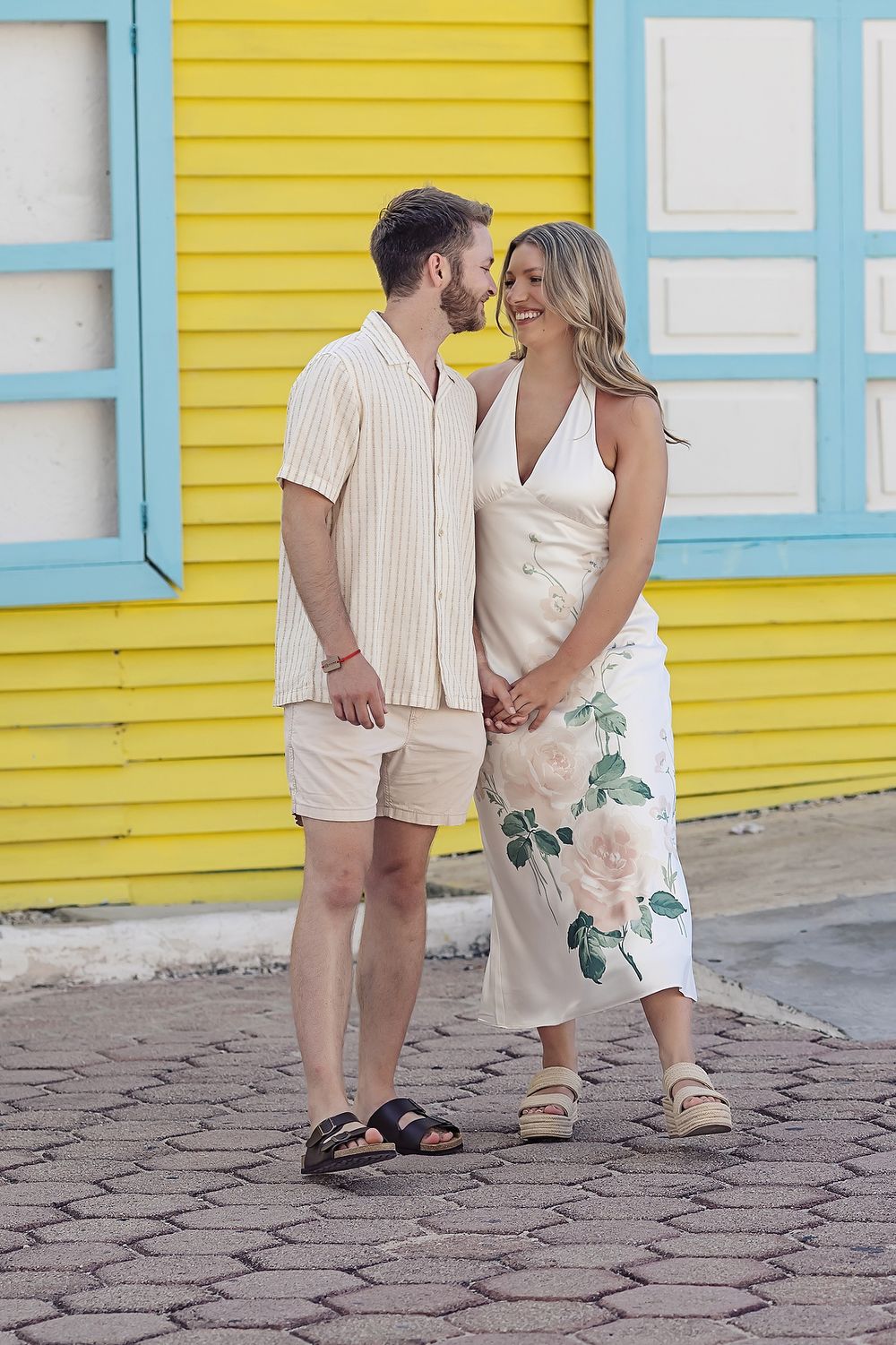 A couple shares a romantic moment in front of a bright yellow wall with blue trim wearing coordinated summer outfits.