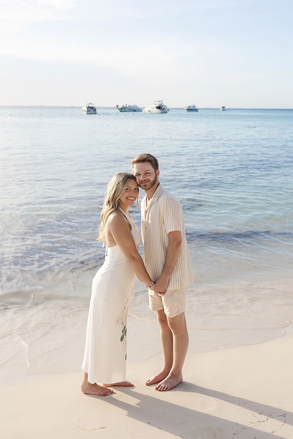 A couple in light colored outfits embraces on a tropical beach with boats visible in the water behind them.
