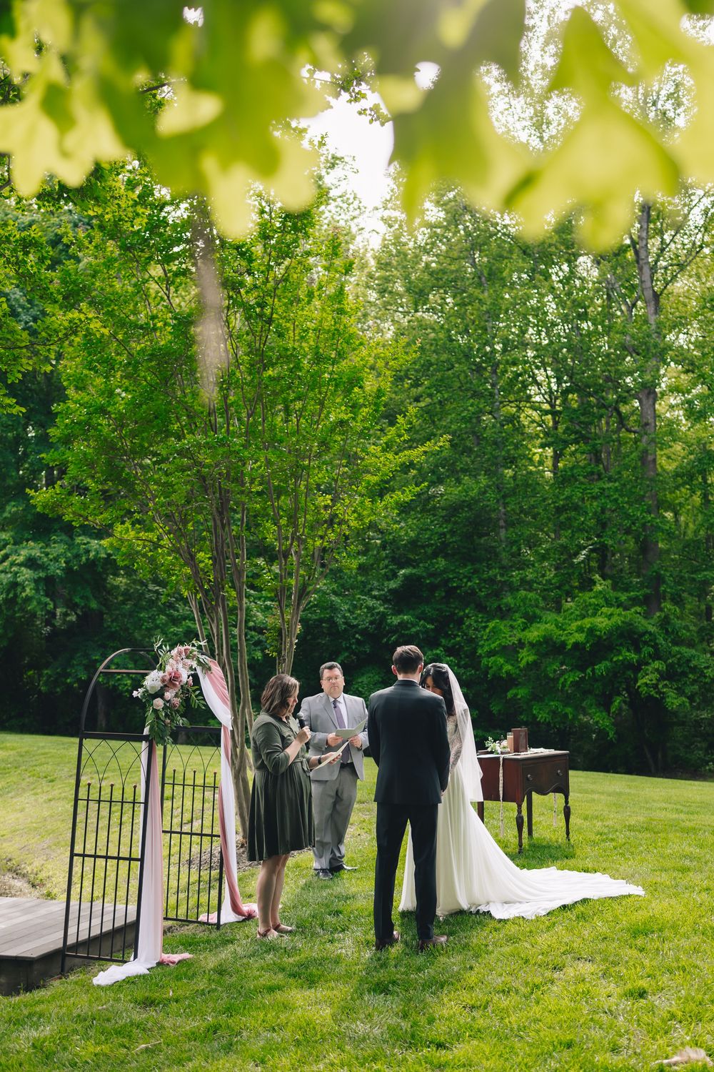 Outdoor wedding ceremony setup with arch and small gathering in lush green garden setting.