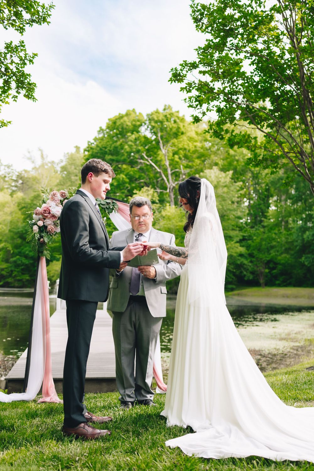 Wedding ceremony taking place outdoors with bride in white gown and minister on lush green lawn.
