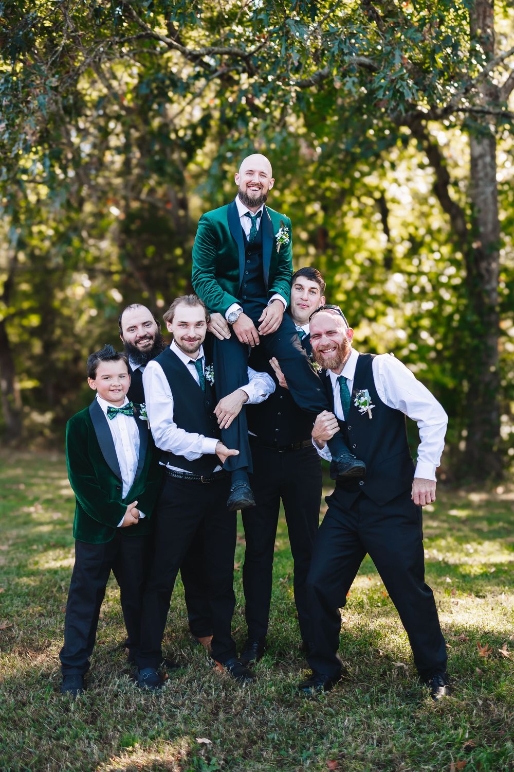 Groomsmen lifting groom in emerald velvet jacket for playful outdoor portrait.