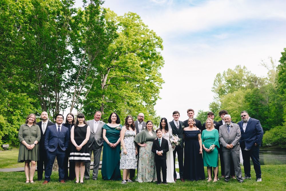 Large wedding party group photo with guests lined up on green lawn during outdoor ceremony.