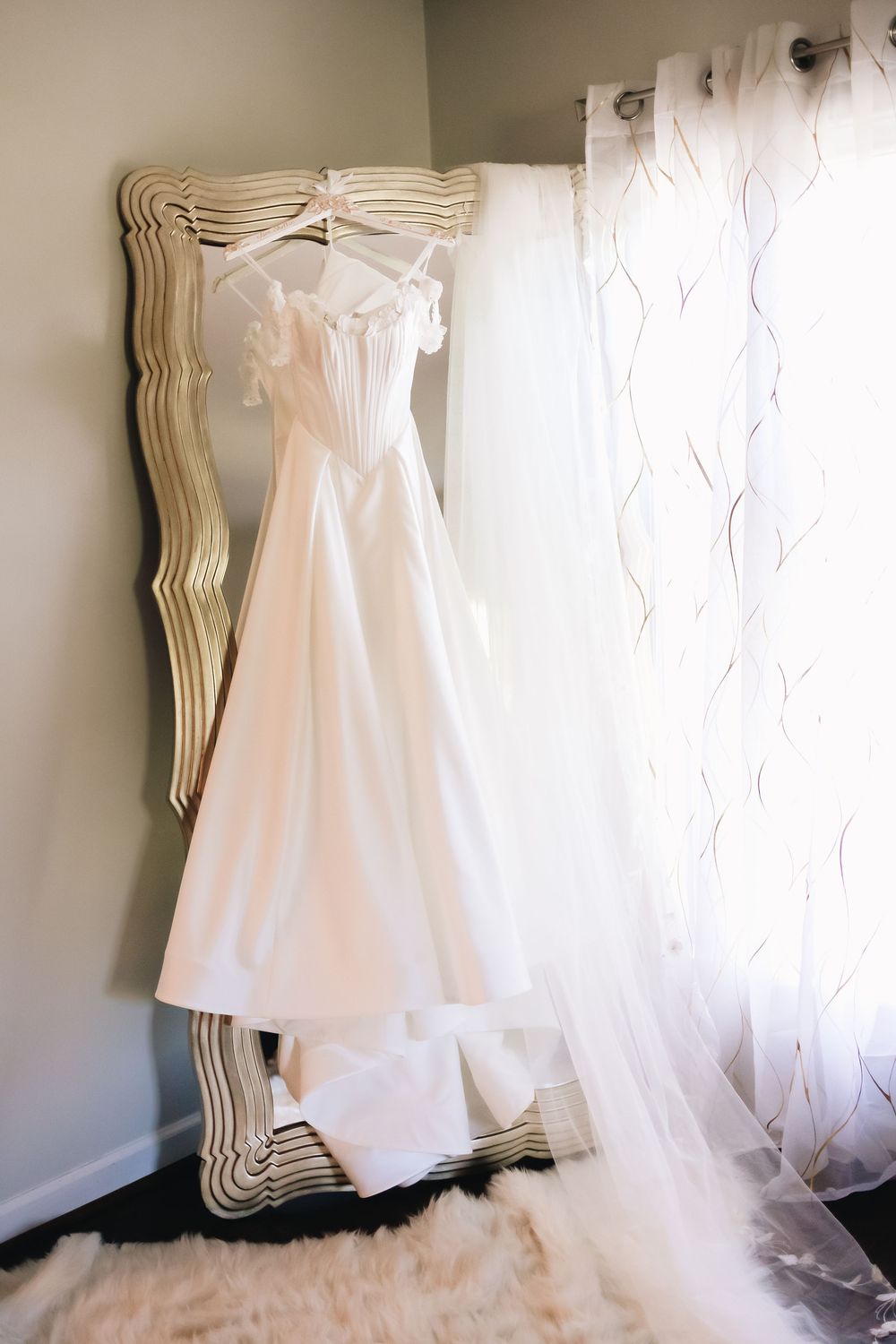 Classic white wedding dress hangs elegantly on decorative mirror with sheer curtains.