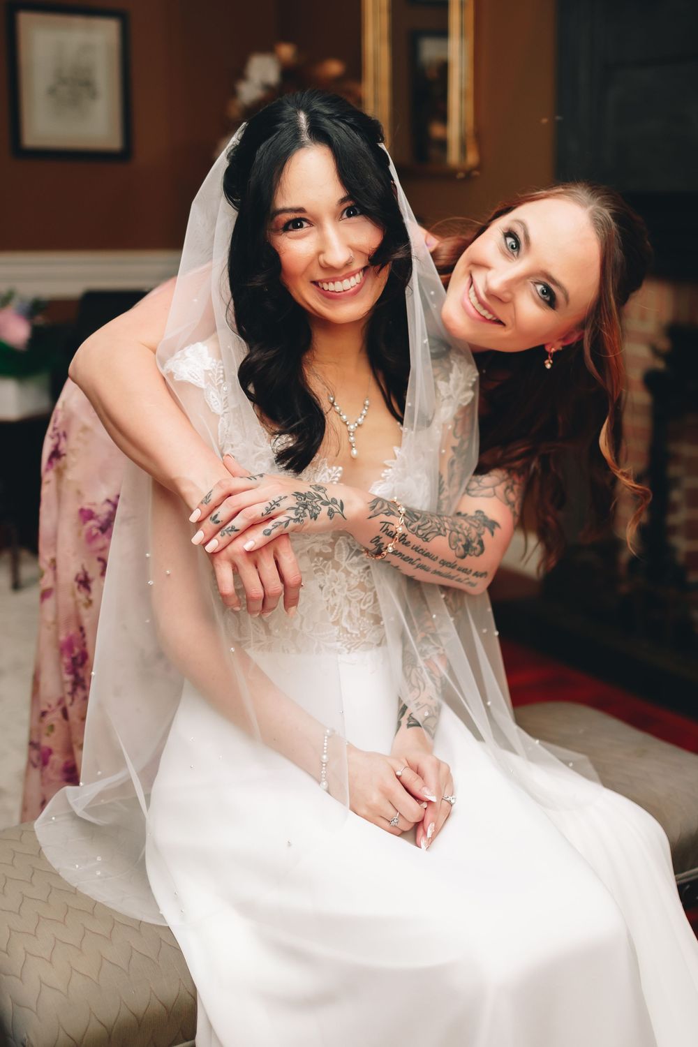 Two women embrace and smile while wearing wedding attire indoors.