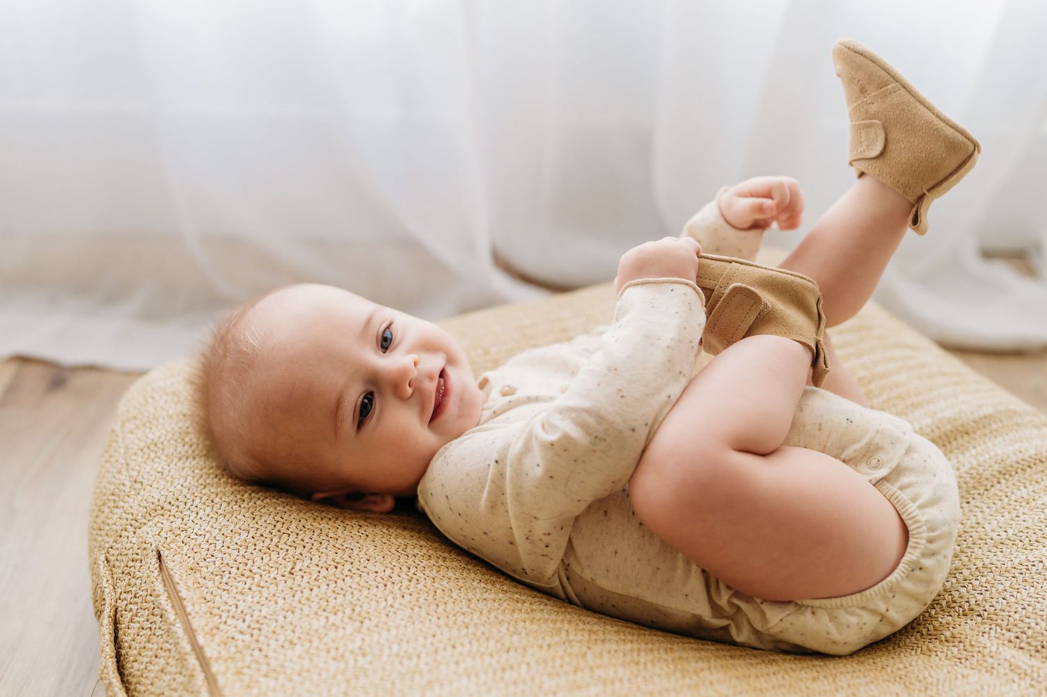Baby lying on cream colored blanket wearing neutral colored outfit in bright indoor setting.