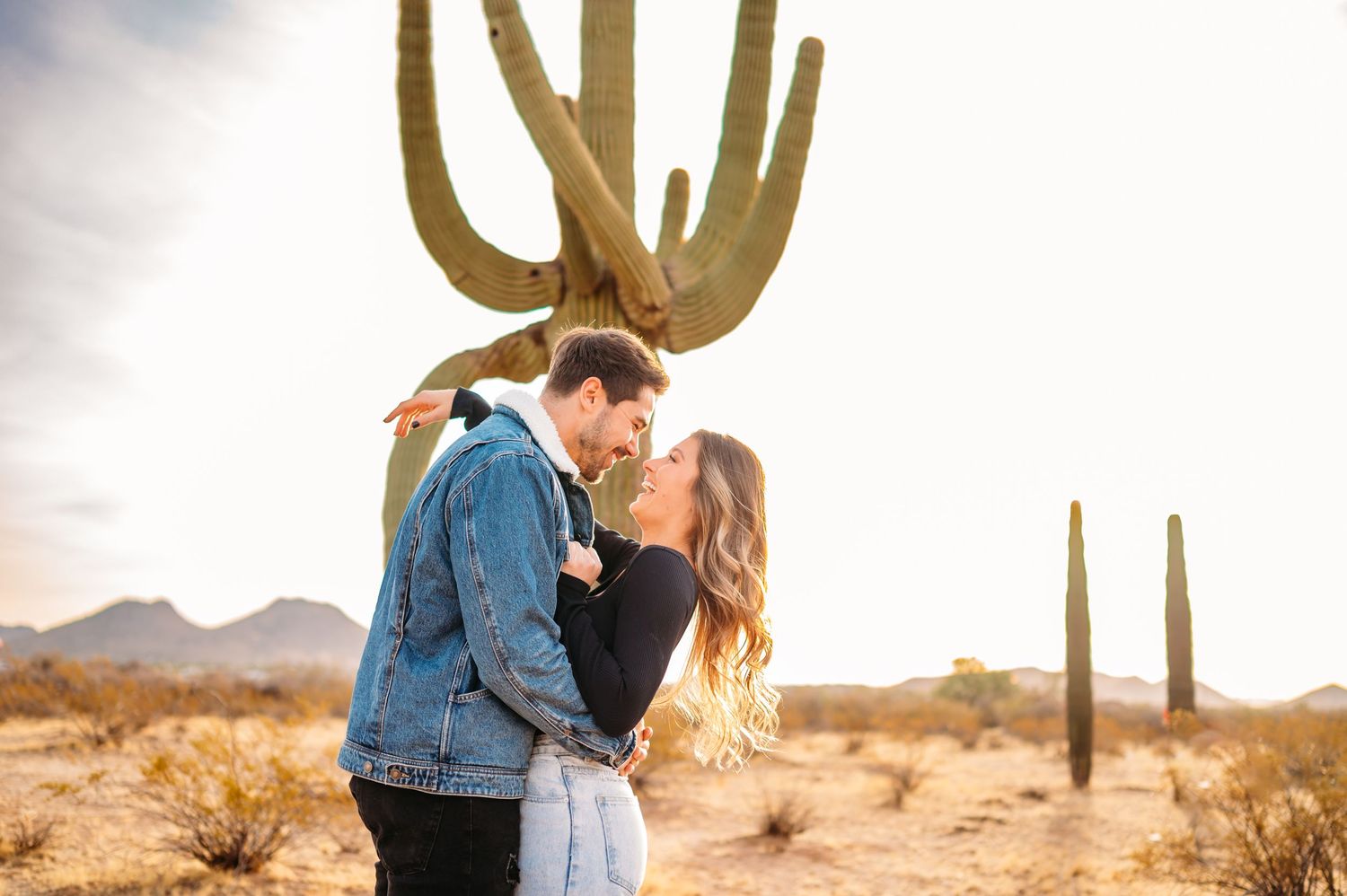 Romantic desert engagement photo with couple kissing beneath towering saguaro cactus.