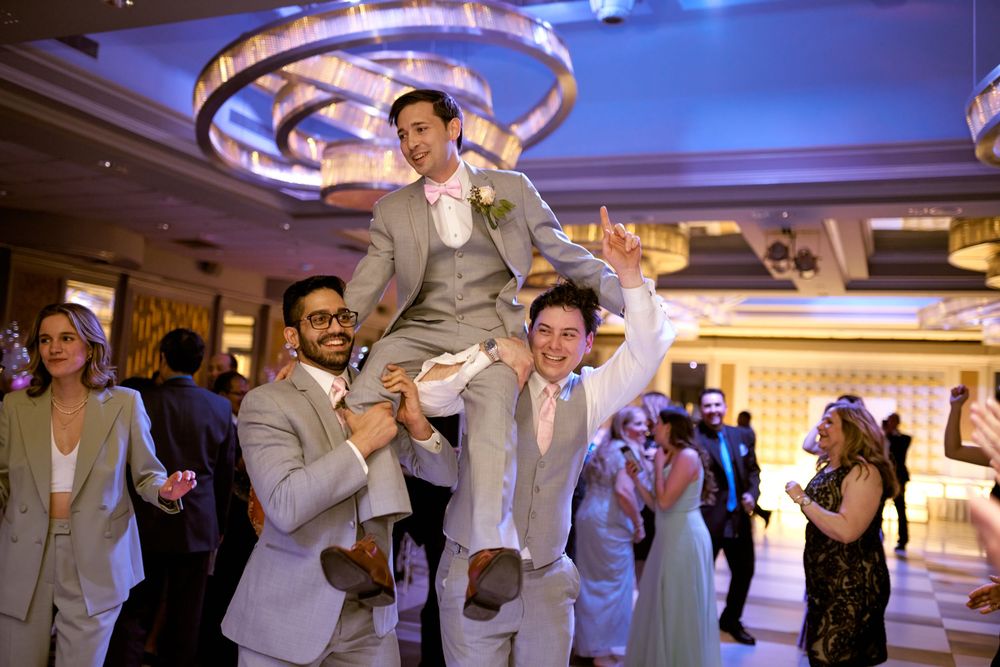 Wedding guests lift someone in grey suit during lively reception dance party.