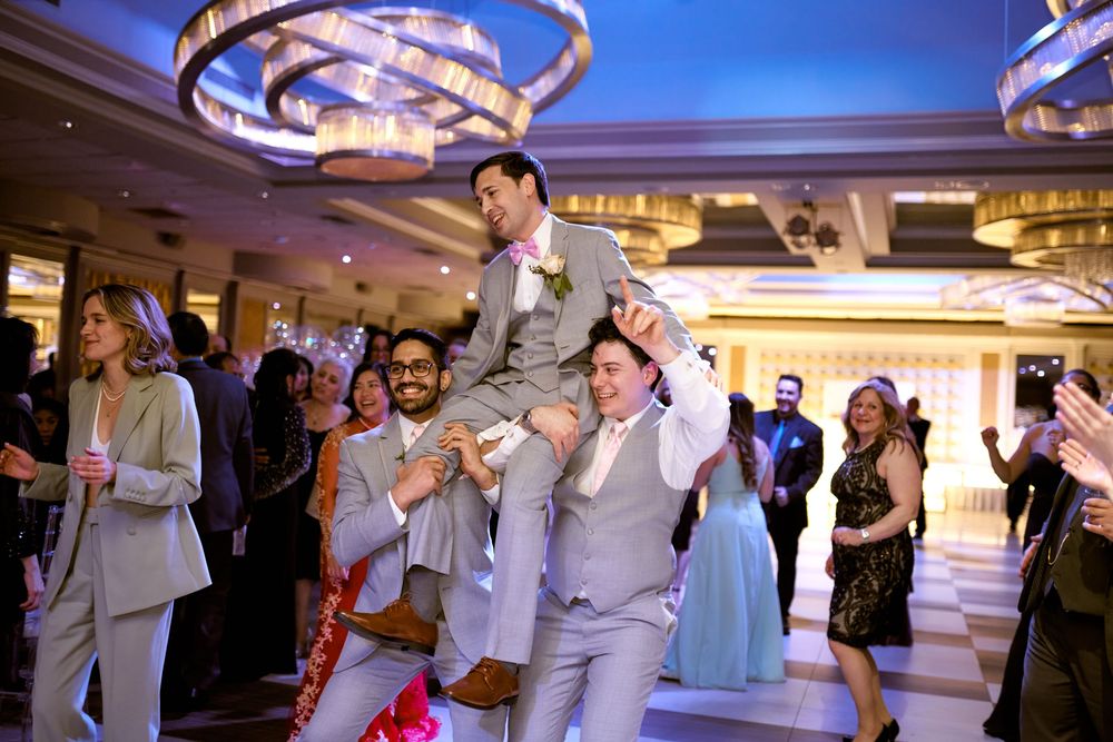 Wedding guests lift and celebrate with someone in a grey suit during reception festivities.