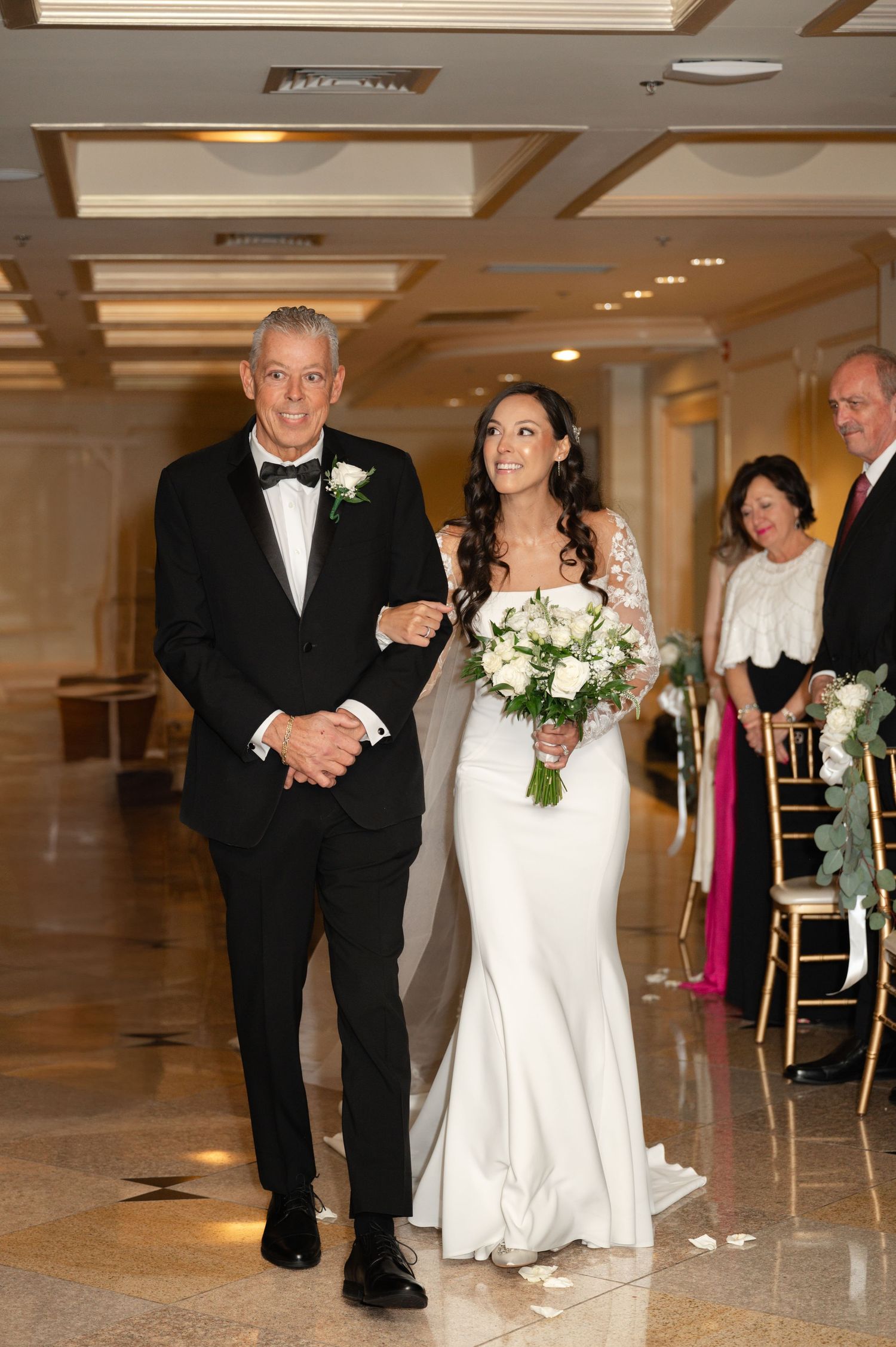 A bride in a fitted white gown walks arm in arm with an escort down an elegant hotel hallway during a wedding ceremony.