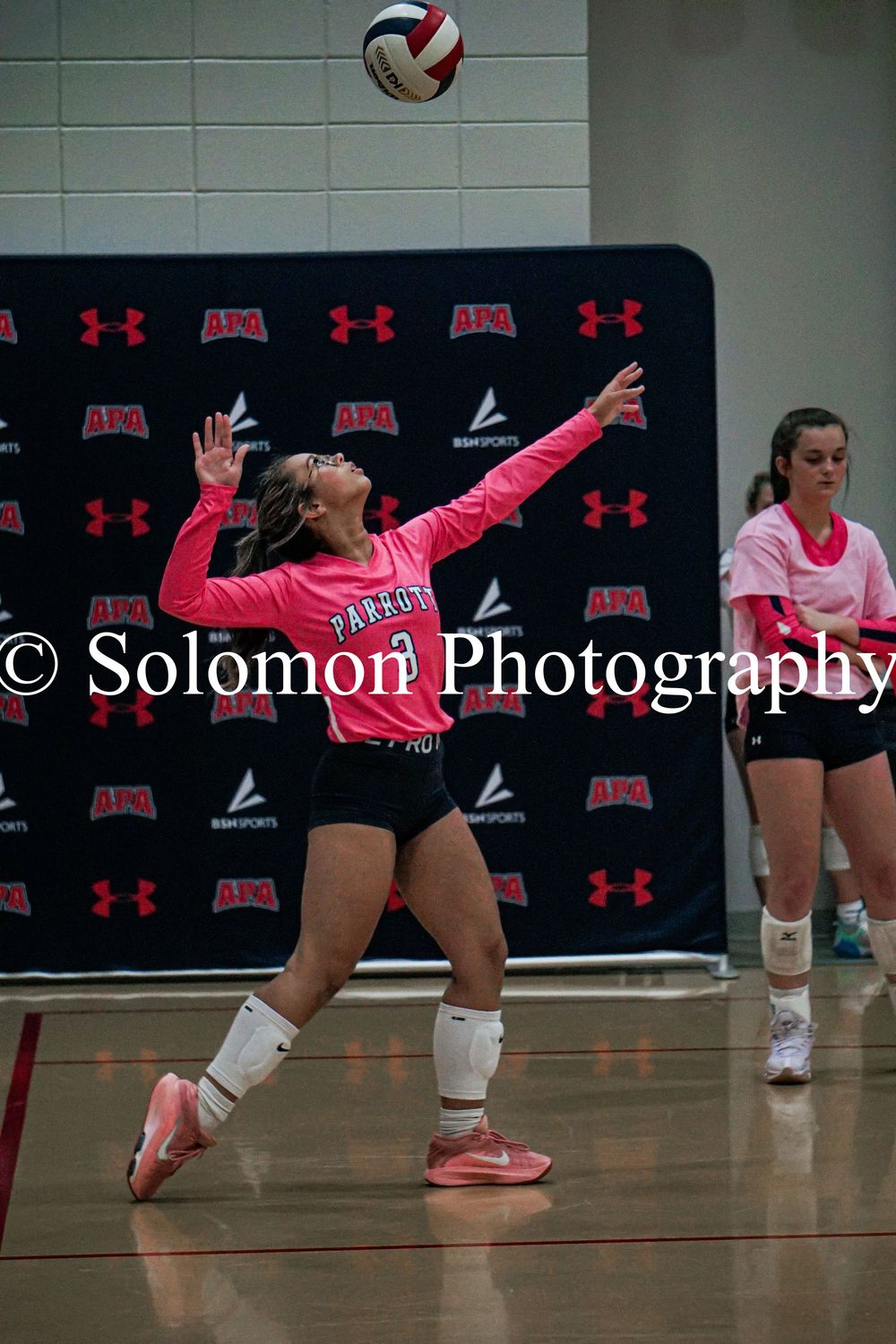 Female volleyball player in pink uniform serves the ball during an indoor court match.