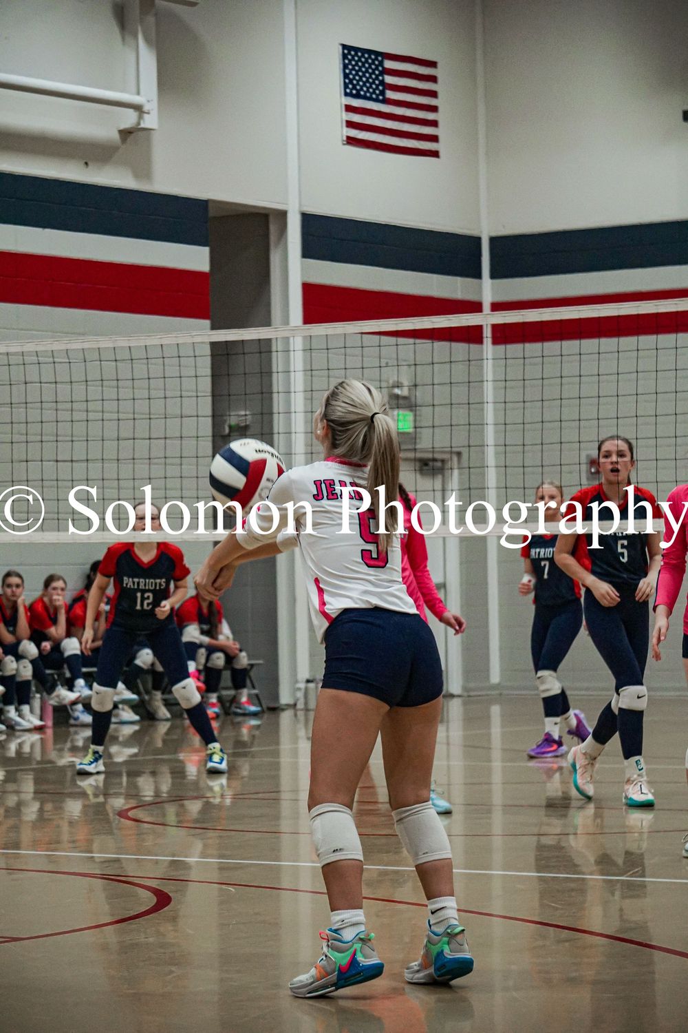 Volleyball player in pink and white uniform prepares to receive the ball near the net.